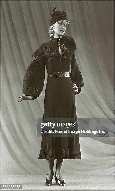 Vintage black and white studio photograph of a full-length mannequin in a svelte tea-length back dress with puffed sleeves, a silver belt, a black...