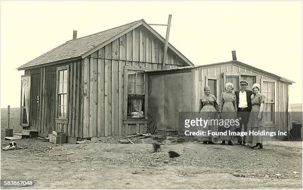 Vintage black and white photograph of a man and three women standing beside small wooden house with no vegetation on a flat prairie, 1890’s. A dog,...