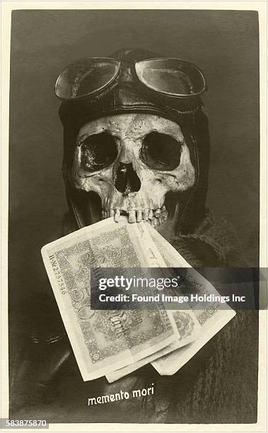 Vintage black and white photograph of a human skull wearing a pilot’s cap, goggles, and a scarf and holding paper money in its teeth in the 1910s,...