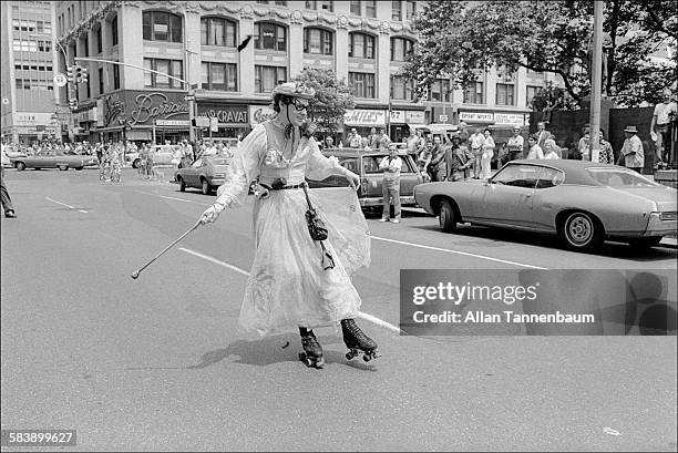 Onlookers watch from the curb on 6th Avenue as a roller-skater in an ankle-length dress, hat, and cat's eye glasses passes them during a Gay Pride...