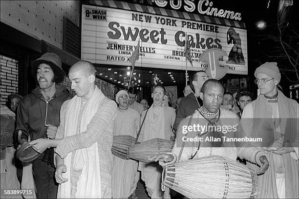 In Times Square, Hare Krishna devotees sing and play percussion instruments as they walk under a cinema marquee that advertises a pornographic movie...