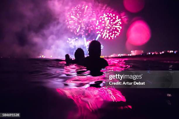 girls having fun enjoying and contemplating the colorful fireworks at night taking a bath in the beach with colorful reflection. - praia noite imagens e fotografias de stock