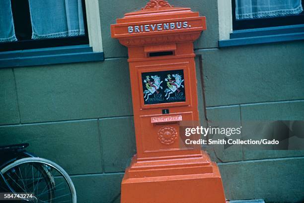 Red mailbox, marked "brievenbus" or "mailbox", with a bicycle leaning against a wall, Netherlands, 1952. .