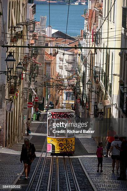 As a local leans out from a window above and others walk uphill, one of the two cars of the funicular railway climbs the steep gradient of on Rua de...