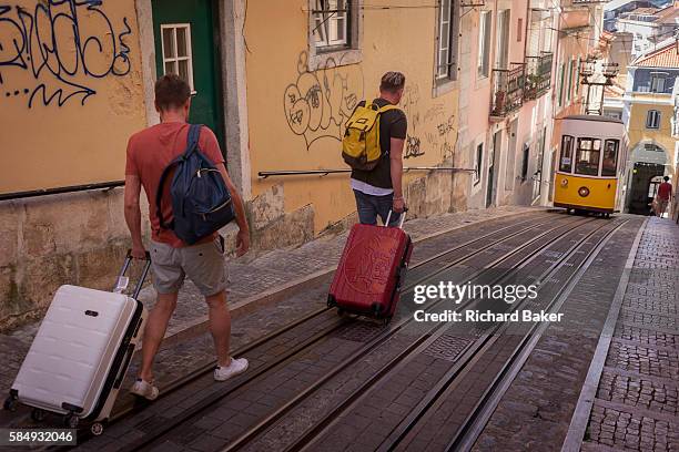 Two tourists walk downhill with their baggage towards one of the two cars of the funicular railway climbing the steep gradient of on Rua de Bica de...