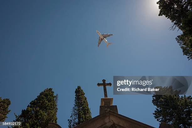 Passing jet airliner flies overhead, above the cross of a family mausoleum, on 14th July 2016, at Prazeres Cemetery, Lisbon, Portugal. Prazeres...