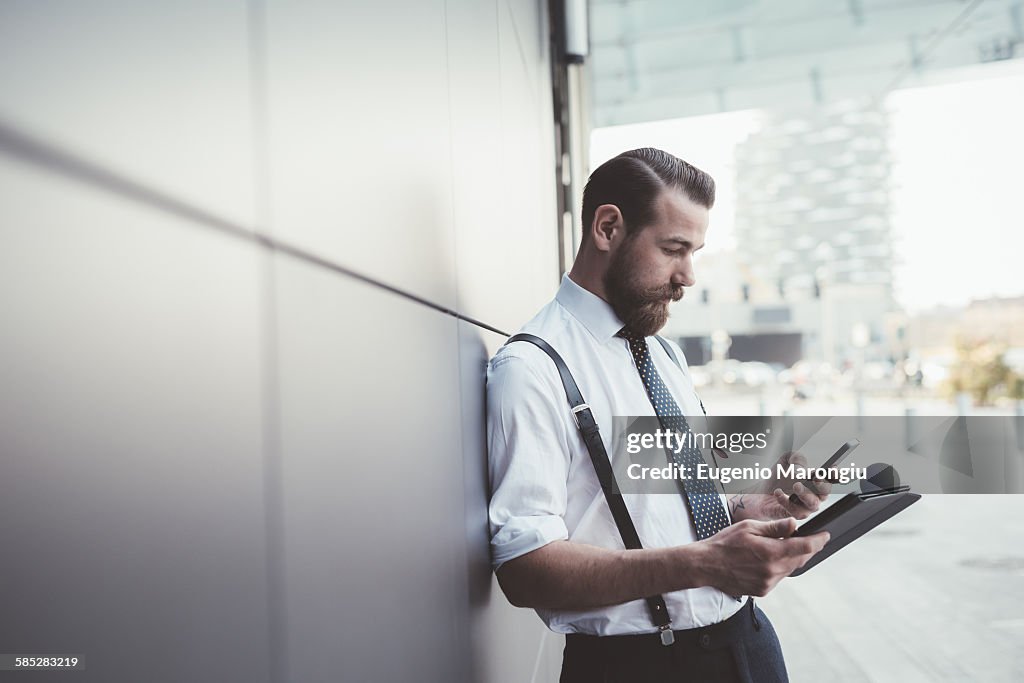 Stylish businessman using smartphone and digital tablet outside office