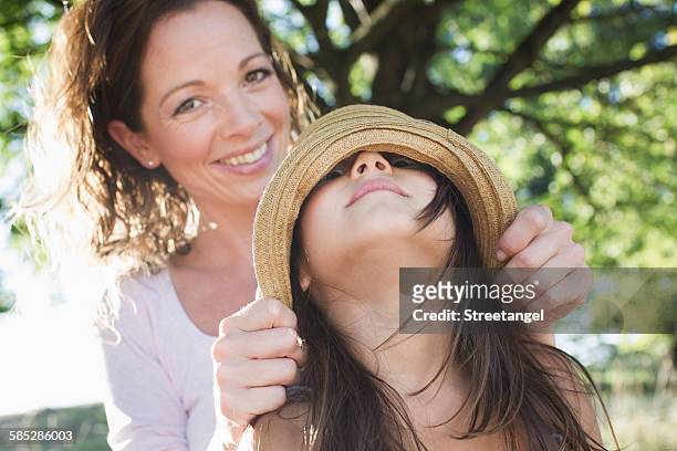 portrait of mature woman covering daughters eyes with straw hat in park - augen zuhalten stock-fotos und bilder