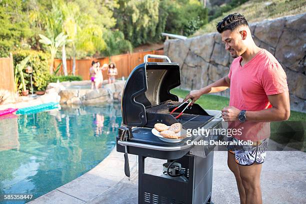 young man cooking burgers on barbecue next to garden swimming pool - 勞動節 北美假日 個照片及圖片檔