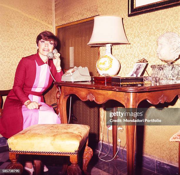 Italian actress Franca Valeri in her house talking on the phone sitting on a bench placed next to a console table. Italy, 1972