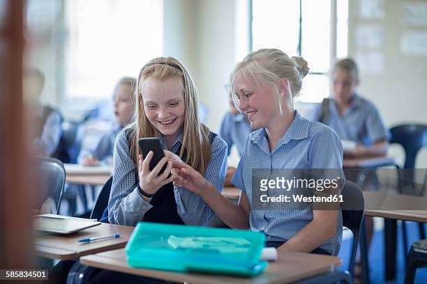 two smiling schoolgirls in classroom looking at cell phone - cell phones in school stock pictures, royalty-free photos & images