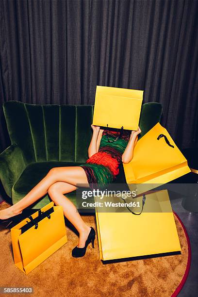 young woman sitting on couch hiding her face behind a shopping bag - zapatos-amarillos fotografías e imágenes de stock