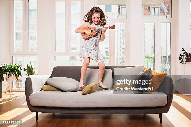 happy girl playing mini guitar while jumping on a couch in living room at home - jump on sofa stock-fotos und bilder