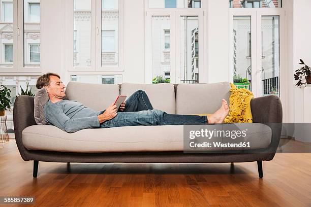 man relaxing with digital tablet on a couch at living room - reclining stock pictures, royalty-free photos & images