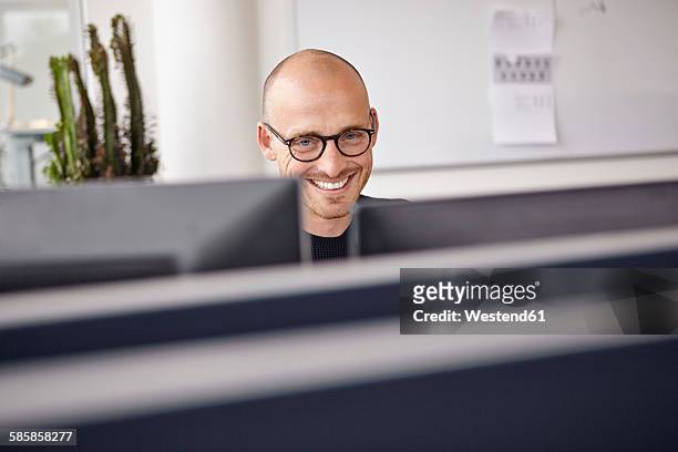 smiling man in office behind computer screens - geheimratsecke stock-fotos und bilder