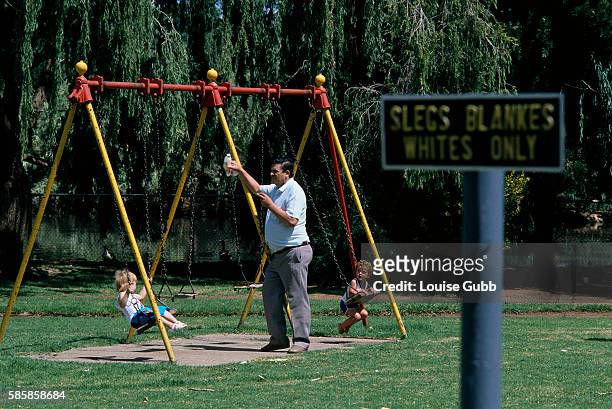 White South African father watches his two young children playing on a swing set near an apartheid sign reading "Slegs-Blankes-Whites Only" in a...