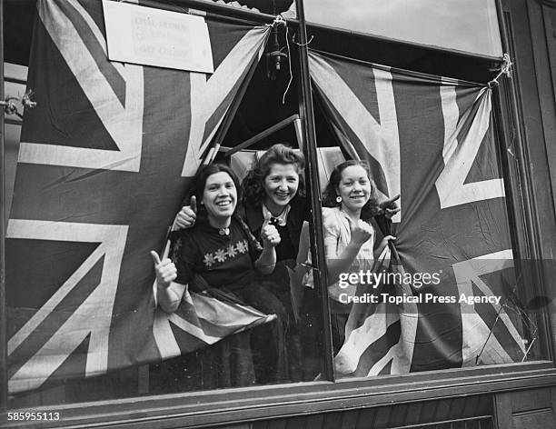 Three young women giving the thumbs-up through union jack flags, which are being used to cover the broken windows of a cycle painting shop in south...