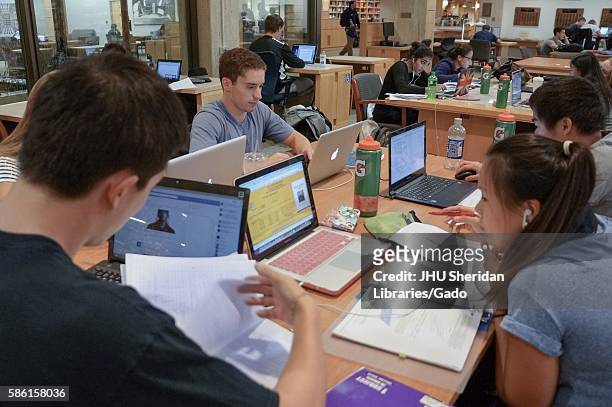 Multiple groups of college students sit together at tables in the Milton S Eisenhower Library at Johns Hopkins University, studying from notebooks...