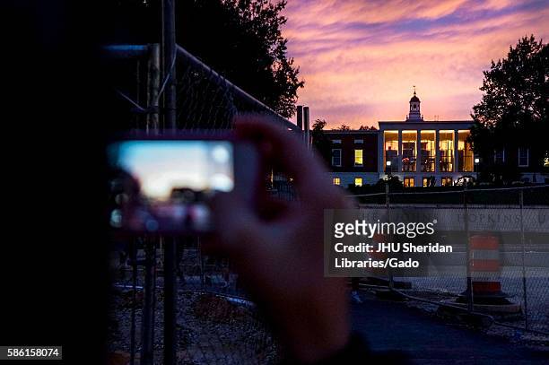 The background of the Johns Hopkins University sign and the Milton S. Eisenhower Library at sunset with a purple and pink clouded sky, with the...