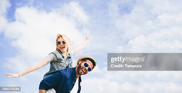 fun piggyback against a cloudy blue sky - couple on beach sunglasses stockfoto's en -beelden