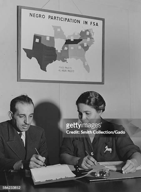 Constance Daniel and Jerome Robinson, Farm Security Administration employees, pose at a desk and look at papers, with a poster in the background...