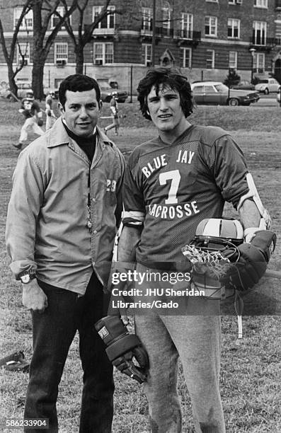 Male lacrosse player for Johns Hopkins University, wearing a number 7 jersey and holding his gear, standing on the field next to head coach Henry...