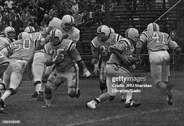 Fans on bleachers in the background watch a football game between two colleges, one of which is Johns Hopkins University, 1970. .