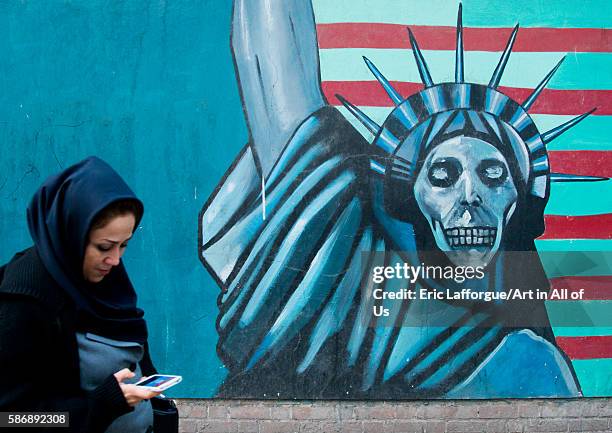 Woman with phone passing in front of an anti-american propoganda slogan depicting statue liberty skeleton on the wall of the united states embassy,...