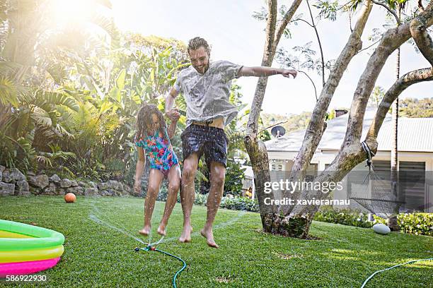 father and daughter jumping in sprinkler at backyard garden - agricultural sprinkler stock pictures, royalty-free photos & images