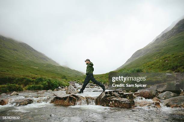 hiker crossing river in mountain valley, glencoe, scotland - hombre mojado fotografías e imágenes de stock