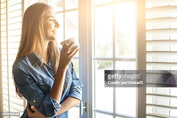 mujer joven bebiendo café y mirando por la ventana - cup day one fotografías e imágenes de stock