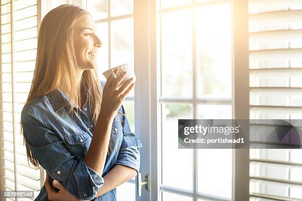 young woman drinking coffee and looking through window - sunlight window stock pictures, royalty-free photos & images