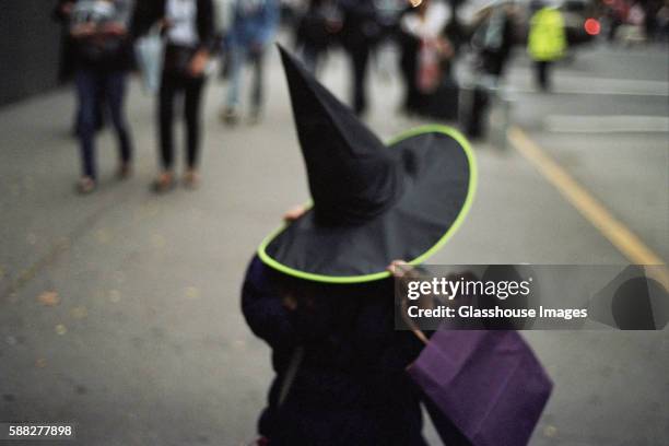 child holding onto witch's hat and purple bag on urban sidewalk, rear view - witch's hat stock pictures, royalty-free photos & images