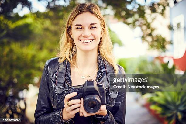portrait of smiling young woman holding slr camera outdoors - spiegelreflexcamera stockfoto's en -beelden
