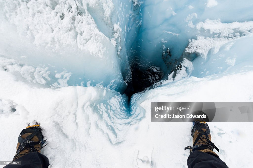 A hiker stands over a moulin, a narrow, tubular chute or crevasse through which water enters a glacier from the surface.