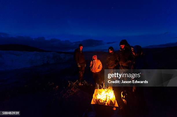 hikers surround a fire to get warm whilst camping at the base of a glacier. - kangerlussuaq stock pictures, royalty-free photos & images