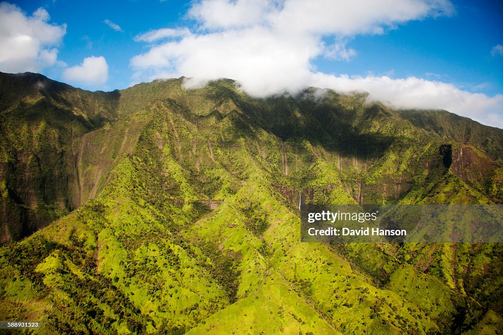 KAUAI, HAWAII, USA. Aerial image of lush green mountain slopes under soft clouds and blue sky.
