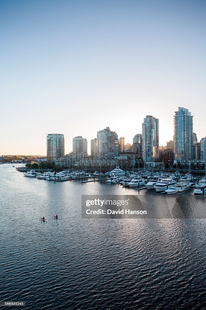 VANCOUVER, BRITISH COLUMBIA, CANADA. Two rowers paddle through calm water with downtown skyline in distance, shot from a bridge.