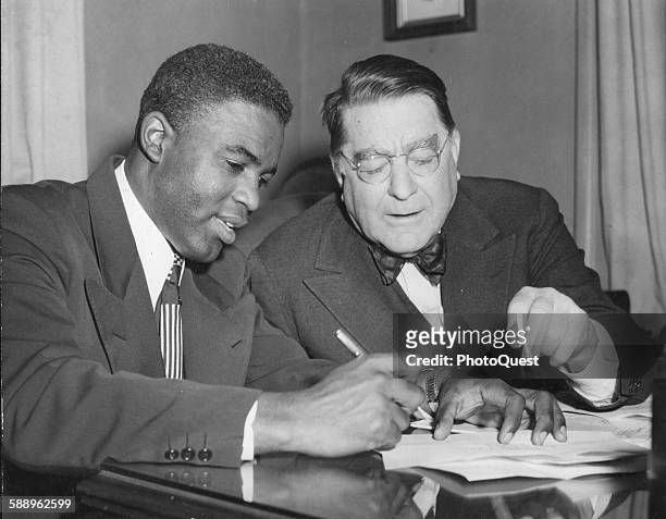 American athlete and second baseman for the Brooklyn Dodgers Jackie Robinson signs his contract for team president Branch Rickey, Brooklyn, New York,...