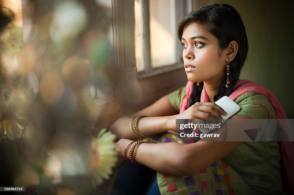 Asian girl next to window with mobile phone looking away.