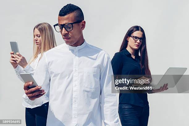 three stylish young people with mini tablet, smartphone and laptop in front of white background - standing with laptop stock-fotos und bilder