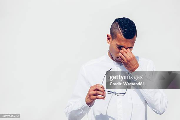young man with shaved hair holding glasses in his hand while rubbing eyes in front of white background - man rubbing eyes stock pictures, royalty-free photos & images