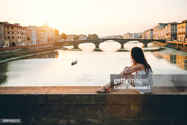 italy, florence, woman wearing white summer dress sitting on a bridge at sunset - florence - italy photos et images de collection