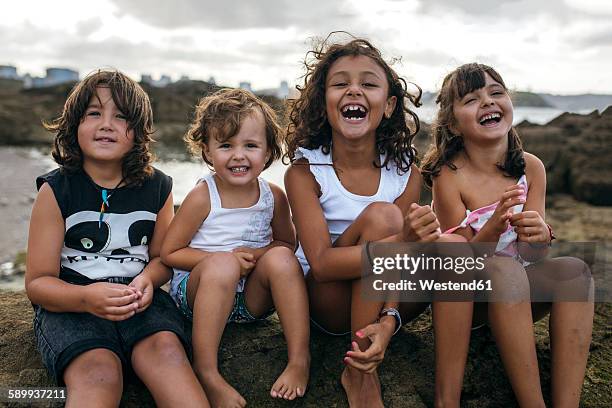 spain, gijon, group picture of four little children sitting at rocky coast having fun - front view portrait of four children sitting on rock stock pictures, royalty-free photos & images