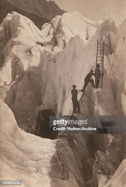 Photographer with his glass plate camera mounted on a tripod straddling a crevasse, and two assistants setting up a ladder, on a glacier in the Alps,...