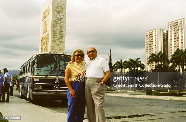 Two tourists stand and pose for a photo, with a Mercedes Benz tour bus in the background, in front of the Monumento de La Nacion A Sus Proceres in...