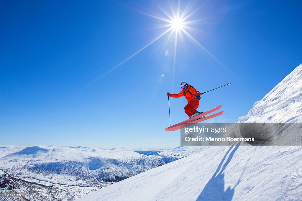 A male freerider in a red suit is jumping from a snow ridge. The sun is shining, the sky is blue.