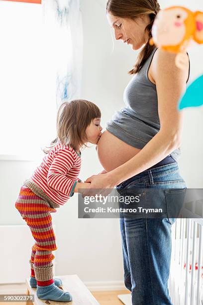 side view of girl kissing on mothers pregnant belly at home - kids belly stock-fotos und bilder