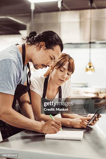 chefs using digital tablet while writing recipe at commercial kitchen counter - digital techniques ストックフォトと画像