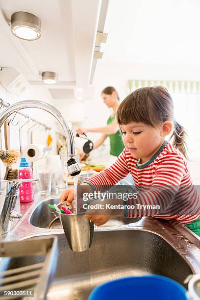 little girl filling water in container with mother in background at kitchen counter - running water faucet stock pictures, royalty-free photos & images